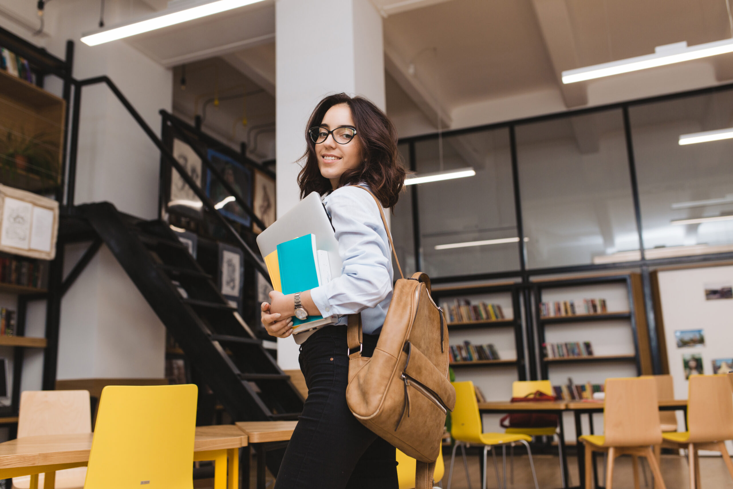 Smiled brunette young woman in black glasses walking with work stuff and laptop in library. Clever student, university life, smiling to camera, cheerful mood